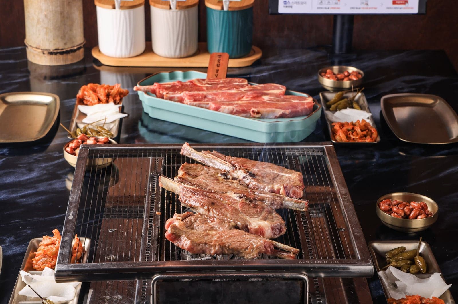 숯불 석쇠 위에서 구워지는 뼈 붙은 양갈비, 뒤쪽 접시에 놓인 생 양갈비와 둘레의 반찬 그릇